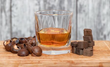 A glass of home-made whiskey sits on a wooden Board . Nearby are the ingredients-fried macadamia shells and burnt oak cubesの写真素材