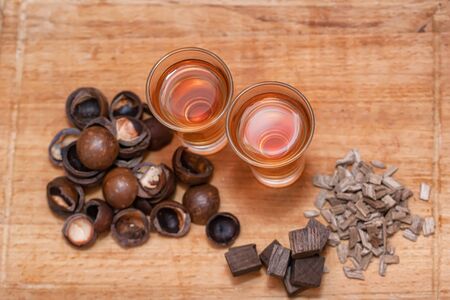 Two shot of home-brewed whiskey stand on a wooden board . Next to it are the ingredients-fried macadamia shells and burnt oak cubesの写真素材