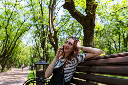 Charming red-haired girl sitting on a bench in a Park and talking on a mobile phoneの写真素材