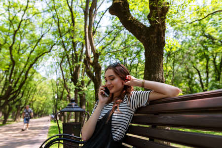 Charming red-haired girl sitting on a bench in a Park and talking on a mobile phoneの写真素材