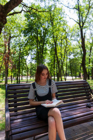 Charming redheaded teen girl in the Park reading a book on a bench and preparing for the examの写真素材