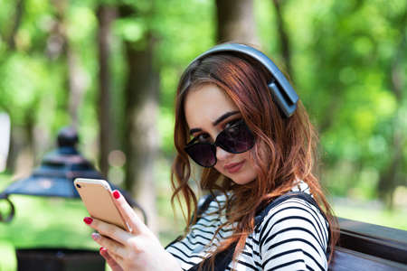 A young pretty red haired girl is listening to music with headphones while sitting on a Park benchの写真素材
