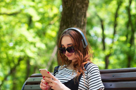 A young pretty red haired girl is listening to music with headphones while sitting on a Park benchの写真素材