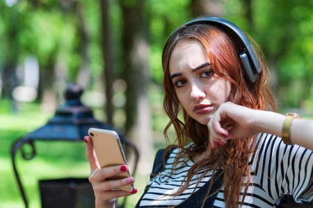 A young pretty red haired girl is listening to music with headphones while sitting on a Park bench. Her look is one of disbeliefの写真素材