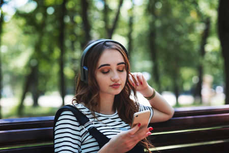 A young pretty red haired girl is listening to music with headphones while sitting on a Park benchの写真素材