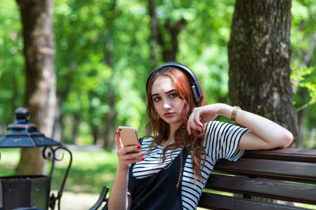 A young pretty red haired girl is listening to music with headphones while sitting on a Park benchの写真素材