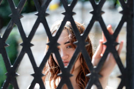 A charming red-haired girl looks at the camera through a forged steel grateの写真素材