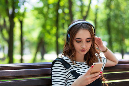 A young pretty red haired girl is listening to music with headphones while sitting on a Park benchの写真素材