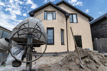 An old concrete mixer, a shovel and a pile of sand against the background of a small cozy cottageの写真素材