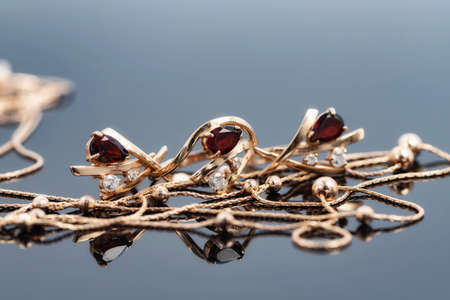 A gold chain necklace with gold beads, a ring and earrings with red rubies lie on a reflective surfaceの写真素材