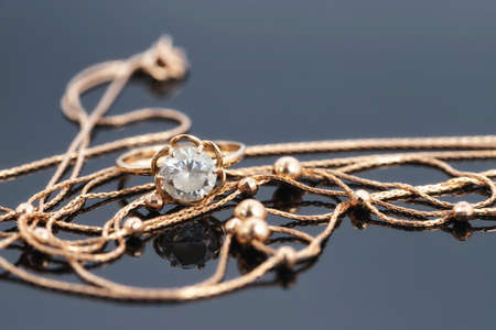 A gold ring with a large gemstone and a gold chain necklace lie on a dark reflective surfaceの写真素材