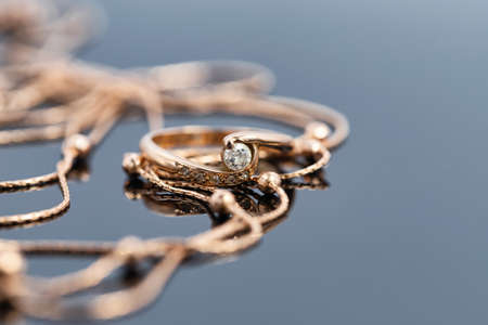 A gold ring with diamond and a gold chain necklace lie on a dark reflective surfaceの写真素材