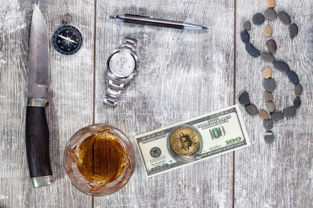 A Damascus steel knife, an elegant handle, a glass of whiskey, a mechanical watch, and a hundred-dollar bill lie on a wooden background. The dollar sign is made of small sea stonesの写真素材
