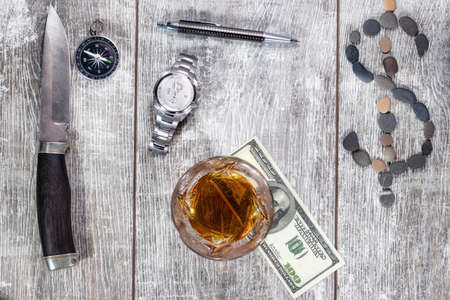 A Damascus steel knife, an elegant handle, a glass of whiskey, a mechanical watch, and a hundred-dollar bill lie on a wooden background. The dollar sign is made of small sea stonesの写真素材