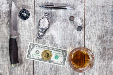 A Damascus steel knife, an elegant handle, a glass of whiskey, a mechanical watch, and a hundred-dollar bill lie on a wooden background. The dollar sign is made of small sea stonesの写真素材