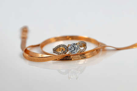 A gold chain with a snake skin weave and a large diamond ring are reflected on the white surface