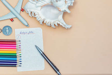A piece of paper from a notebook with a plan for today, colored pencils, a compass, a seashell and a toy airplane on a beige backgroundの写真素材