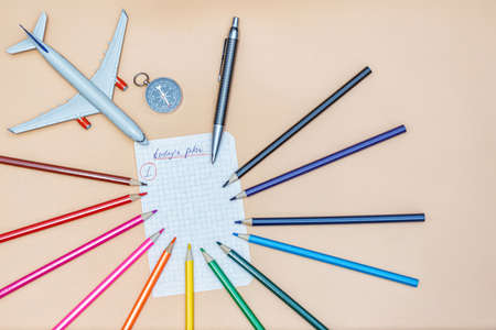A piece of paper from a notebook with a plan for today, colored pencils, a compass, a seashell and a toy airplane on a beige backgroundの写真素材