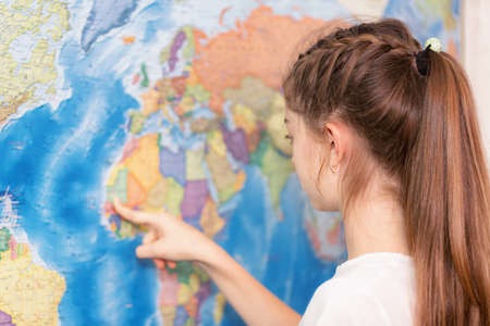 A school student studies geography on a large map of the world on the wall. She points her finger at...
