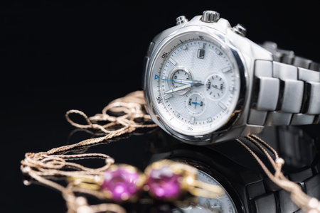 Solid men's watch with a chronometer in a titanium case lies against the background of gold women's jewelry on a black reflective surfaceの写真素材