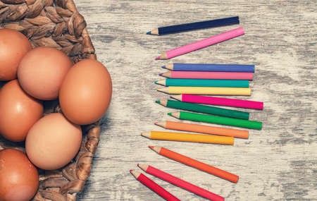 Unpainted beige chicken eggs and multicolored pencils lie on a wooden countertop.の写真素材