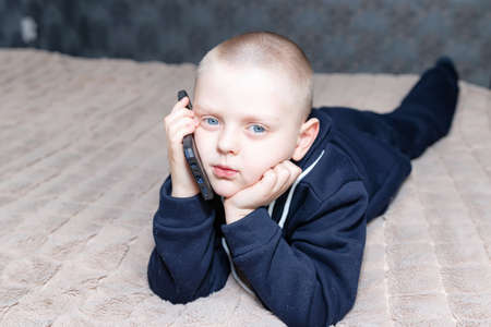 A preschool boy talks on the phone lying on the bedの写真素材