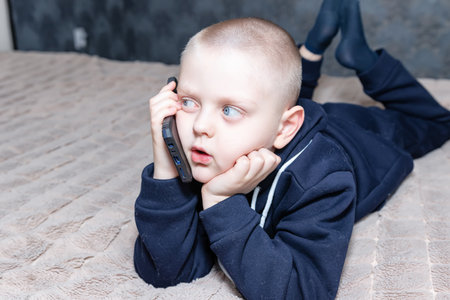 A preschool boy talks on the phone lying on the bedの写真素材