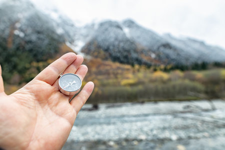 A hand holds a compass against the background of snow-capped mountains and a river, symbolizing the search for a path and harmony with natureの写真素材