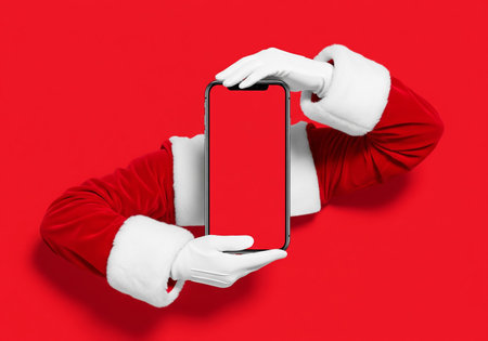 Santa Claus holding a smartphone with a blank screen on a red backgroundの素材