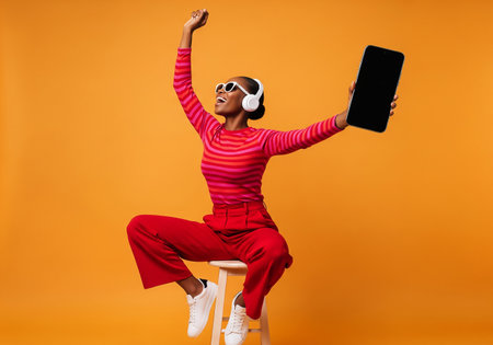 Cheerful african american woman in red pants and t-shirt sitting on chair and showing smartphone with blank screen over yellow backgroundの素材