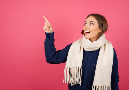 Portrait of a beautiful young woman in winter clothes pointing away on a pink backgroundの素材