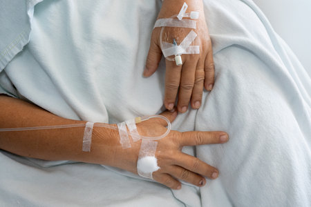 Hand of patient lying on bed in ward at hospital with saline solution tube injection and sterile...