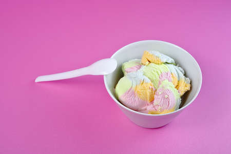 White bowl with colored ice cream and a plastic spoon on a pink background.