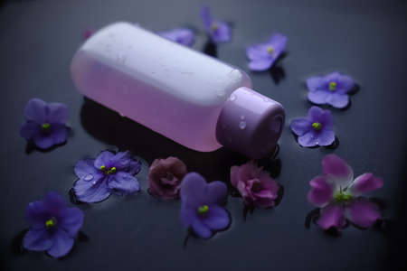 A jar of face tonic on a dark grey background with water and purple violetsの写真素材