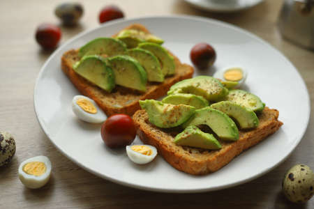 Toast with sliced avocado quail eggs cherry tomatoes on a white plate close-up. Healthy breakfast.