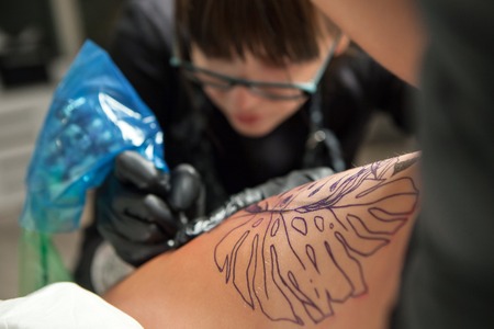 Young cute girl tattoo master at work on patterning on the model's body. Concentration and focus on...