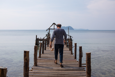 An old man in a striped t-shirt with a cap in his hand and a bald head walks along the pier to the sea, the view from the backの写真素材
