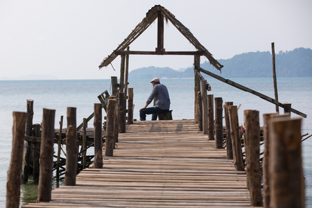 Sad old man in a cap sitting on the pier and looking at the sea, the view from the backの写真素材