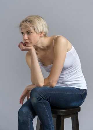 Pensive posing studio photoshoot, woman sitting hunched over. Thoughtful mood, model with shirt blond hair in casual outfit on bar stool portrait shotの写真素材