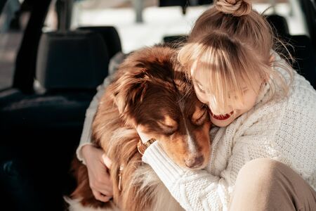 Beautiful woman sit and cuddle in the trunk of a minivan with a dog breed Lassie Australian Shepherd. Caring for a dog. Dog is a mans friend, a true friend. Sunny winter day.Close-upの写真素材