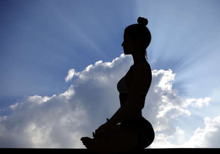 Woman side silhouette in Yoga Easy Meditating pose with rays of sunlight through Cloudy sky in backgroundの写真素材