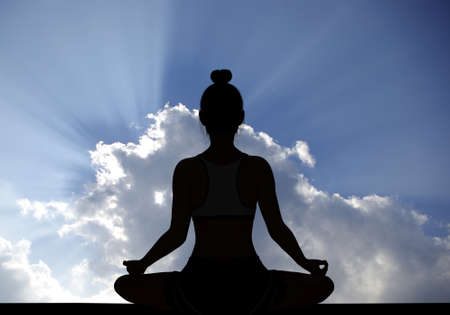 Woman silhouette in Yoga Easy Meditating pose with rays of sunlight through Cloudy sky in backgroundの写真素材