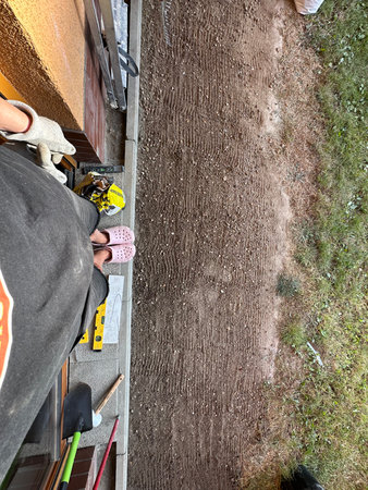 Individual in pink slippers and gloves stands on freshly tilled dirt, surrounded by gardening tools, highlighting the process of outdoor landscaping and home improvementの写真素材