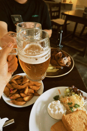Friends raise beer mugs in celebration at a restaurant, surrounded by plates of fries, salad, and grilled meat, capturing a joyful dining experienceの写真素材