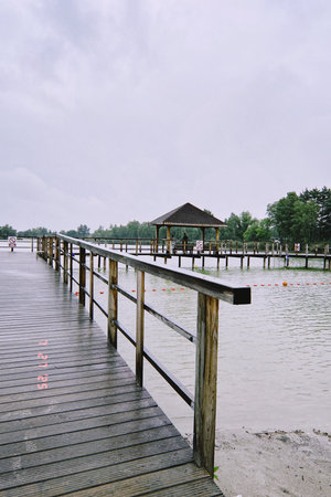 Scenic wooden pier stretches over tranquil water, leading to a gazebo surrounded by greenery, under a cloudy sky, evoking a peaceful outdoor ambianceの写真素材