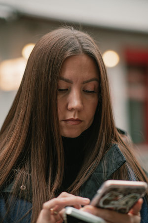 Female with long brown hair is intently using smartphone outdoors, wearing a stylish black jacket, with soft blurred lights creating a vibrant atmosphereの写真素材