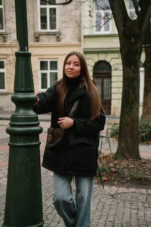 Female with brown hair, dressed in black jacket and jeans, poses near green lamppost in city environment, reflecting a relaxed urban atmosphereの写真素材