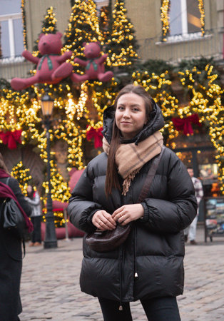Lwiw, Ukraine - November 28, 2025: Young woman wearing a black puffer jacket poses in a beautifully decorated square with Christmas lights and teddy bearsのeditorial素材