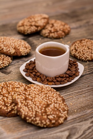 Small white cup of coffee, roasted coffee beans, cookies with sesame seeds on wooden backgroundの写真素材