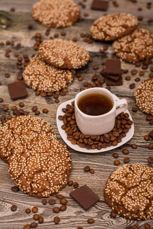 Small white cup of coffee, roasted coffee beans, cookies with sesame seeds, slices of chocolate on wooden backgroundの写真素材